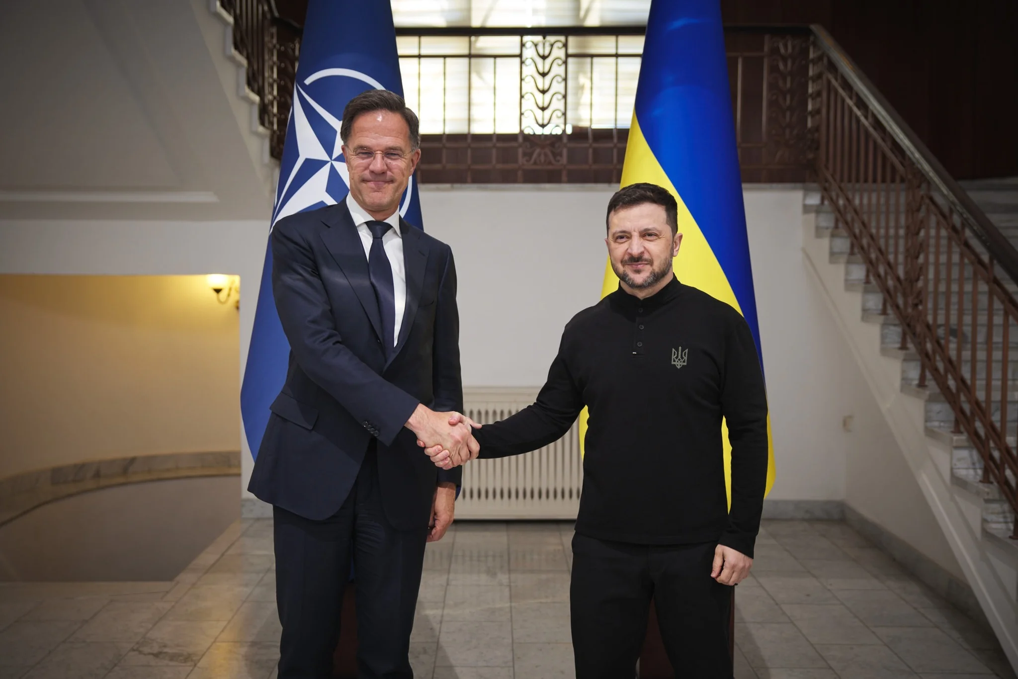 Зеленский: В НАТО выросла поддержка членства Украины Зеленский пожимает руку представителю НАТО на фоне флагов Альянса и Украины